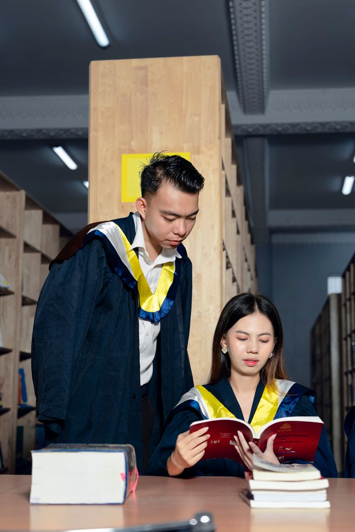 Two students in graduation gowns studying together in a library setting.