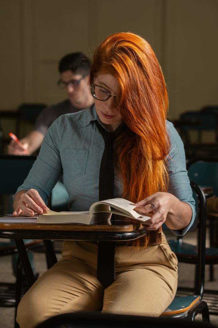 Services A student with glasses and red hair attentively reading a book in a classroom setting.