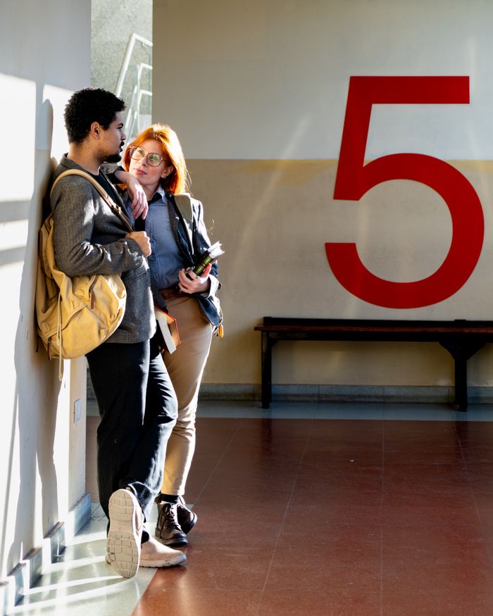 Services Two students discussing in a sunlit hallway in Buenos Aires, leaning against a wall marked '5'.