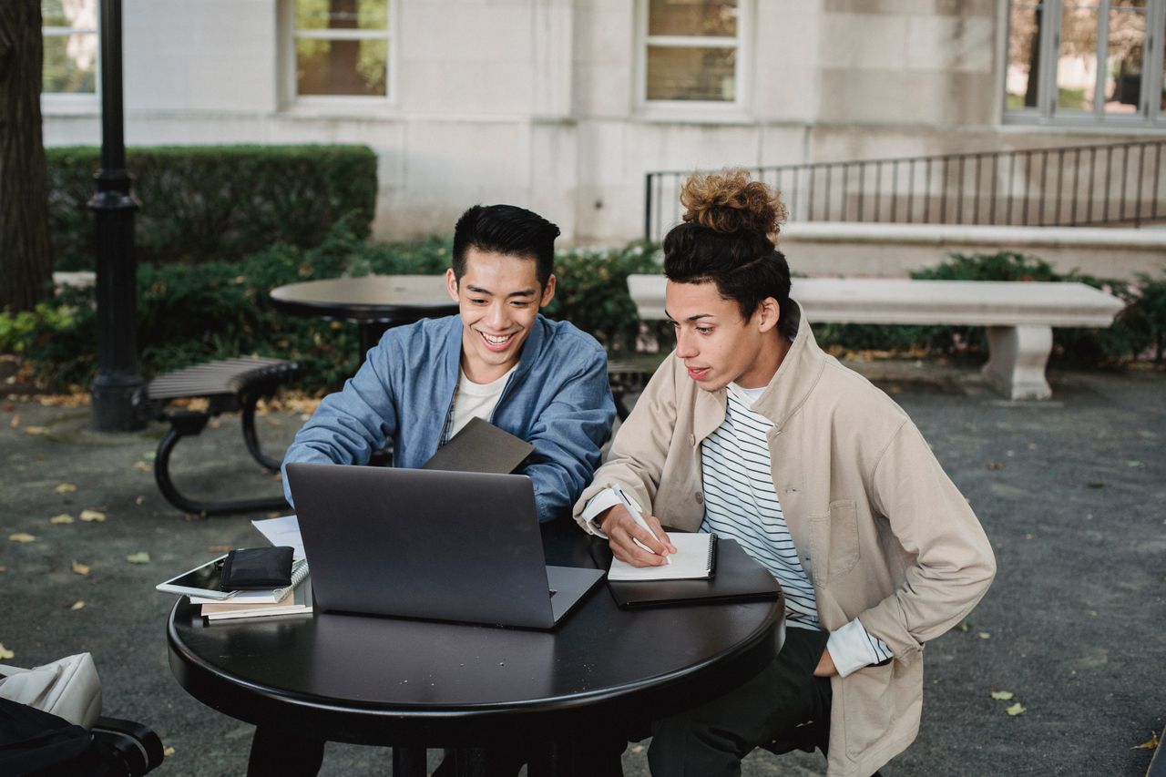 Smiling multiracial male classmates sitting at table and browsing netbook while taking notes in notebook