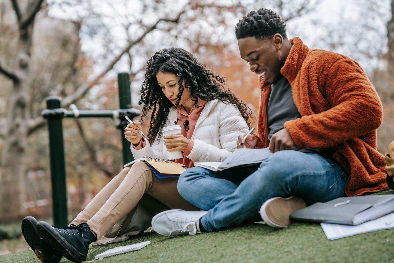From below full body of ethnic woman and black man with takeaway coffee taking notes in notebooks while studying together on street